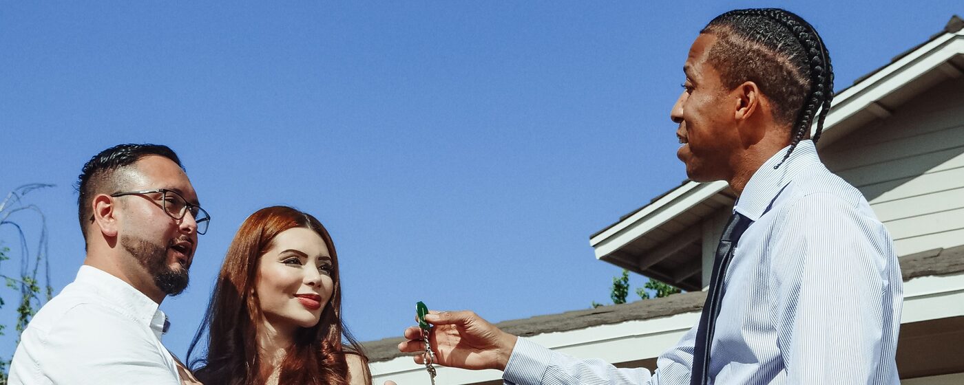 Real estate agent handing house keys to buyers in front of a sold property sign